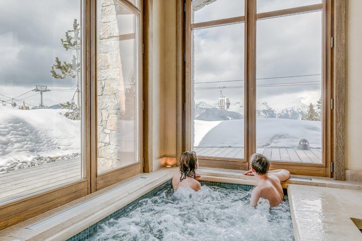 Two people in a hot tub, enjoying a snowy mountain view with a ski lift through large windows.