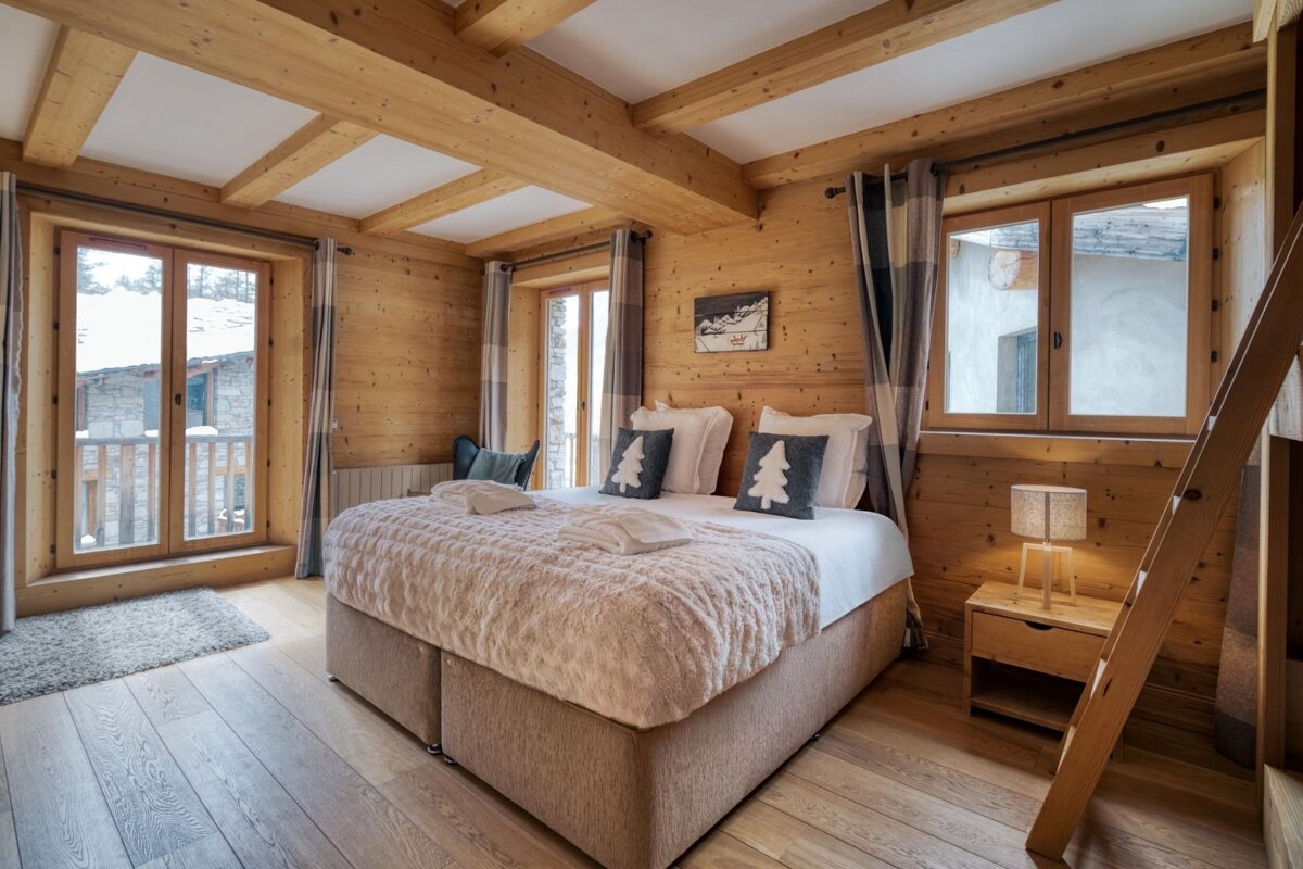 Cozy, wood-paneled bedroom with a large bed, festive tree pillows, and windows, creating a warm, inviting cabin-like ambiance.