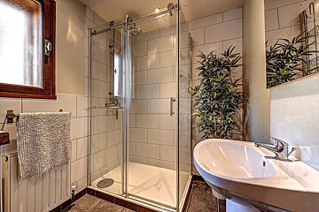 A bright bathroom features a glass shower, white pedestal sink, bamboo plant, and white tiled walls. A wooden-framed window and towel rack are also visible.