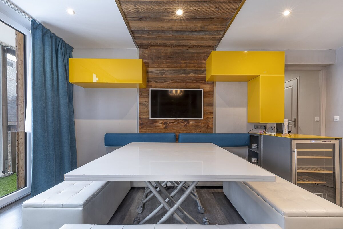 Modern dining area featuring a large white table, white and blue seating, yellow overhead cabinets, and a rustic wood accent TV wall.