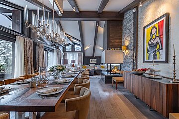 Luxurious chalet interior featuring a long dining table, stylish chandelier, fireplace, and rustic wooden beams.