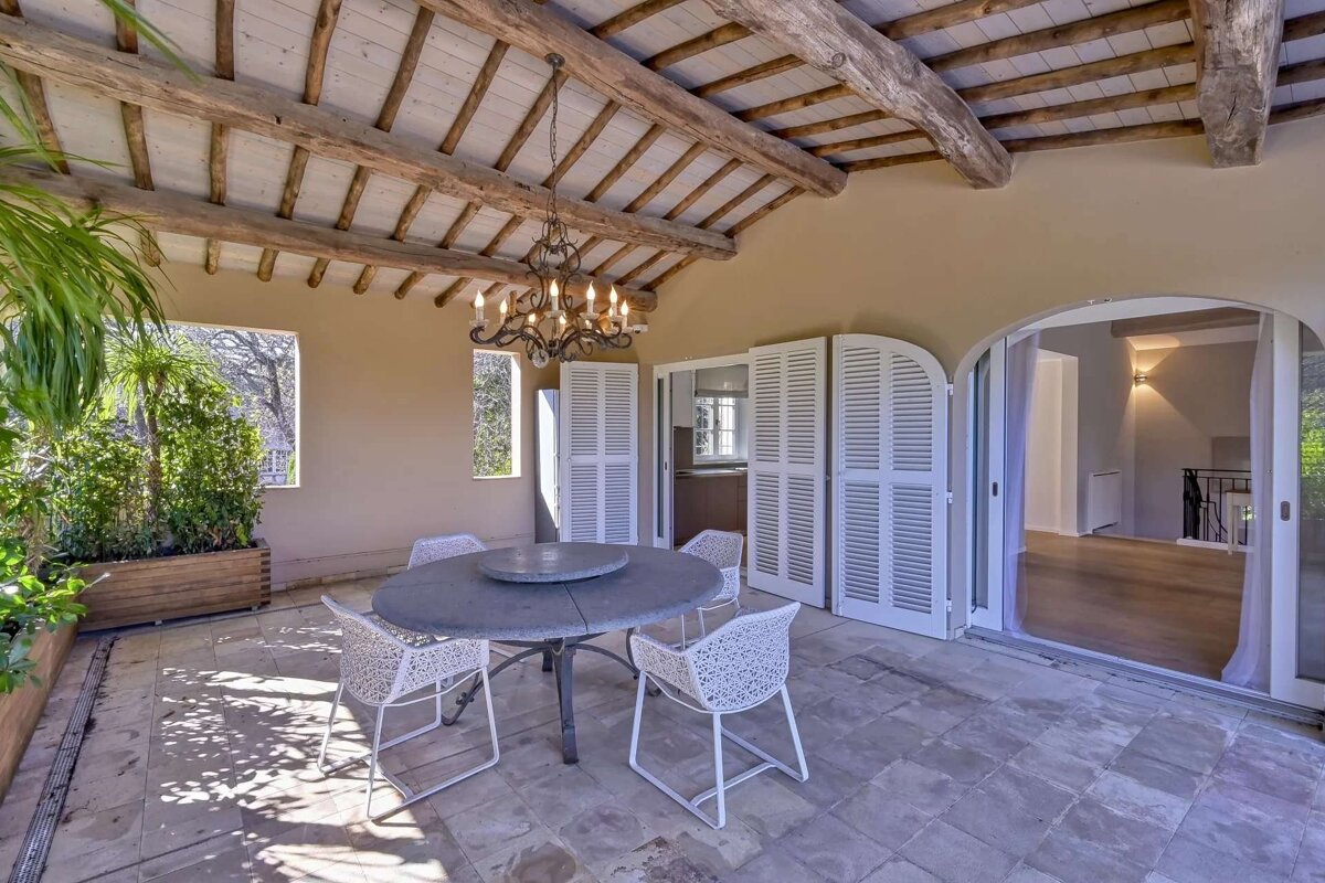 A rustic covered patio with exposed wooden beams, a chandelier, dining set, potted plants, and arched openings leading to an interior.