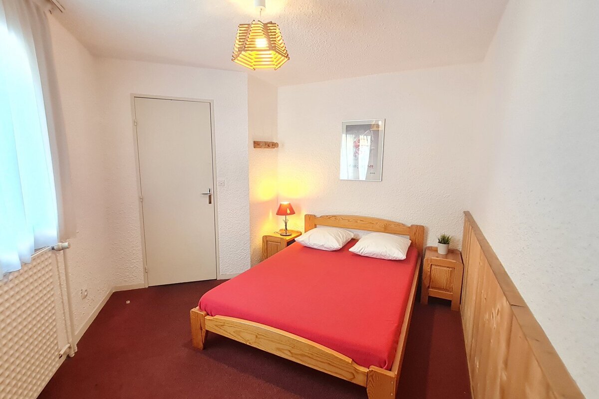 A simple bedroom with a wooden double bed, red bedding, white walls, and dark red carpet. It features nightstands, lamps, a picture, and a window.