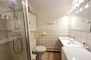 A small, functional bathroom with a glass shower, toilet, sink, and mirror. It features white tiled walls and light brown floor tiles.