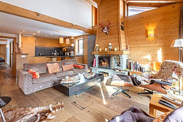 Cozy, wooden alpine chalet living room with a stone fireplace, comfy sofa, lounge chair, and an open kitchen in the background.
