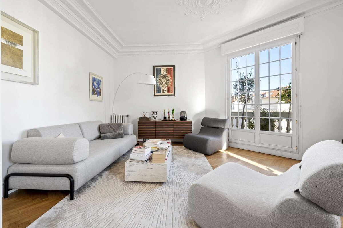 A bright, modern living room featuring light gray sofa & chairs, white walls, wood floor, large window, and ornate ceiling details.