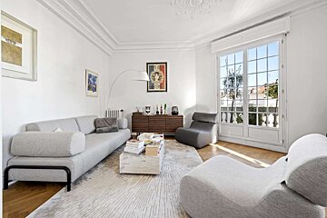 A modern, sunlit living room with light gray sofa, curvy chairs, a rectangular coffee table, and a large window. White walls and wooden floor.