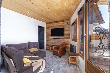 Cozy chalet living room with wood-paneled walls/ceiling, a gray sectional, TV, and leather chair. Glass doors open to a balcony with mountain views.