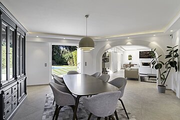 A modern dining room with a sleek table and gray chairs opens to a lush backyard with a pool, connecting to a bright living room via white arches.