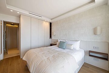 Modern, minimalist bedroom with white double bed, built-in wardrobe, wooden floor, and visible en-suite shower.