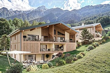Modern alpine residences with wood accents and balconies are nestled on a green, terraced hillside, backed by stunning, snow-capped mountains under a blue sky.