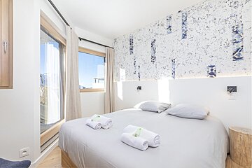 Modern, bright bedroom featuring a double bed with rolled towels, abstract patterned wallpaper, and a window showing a blue sky.