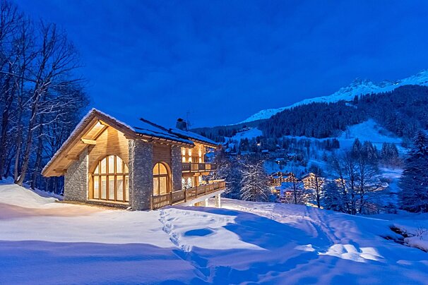 An illuminated, luxurious wooden & stone lodge nestled in deep snow with glowing windows, set against a dark blue sky and snow-capped mountains.