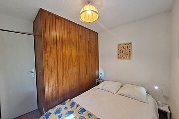 A cozy bedroom with a bed, white walls, and a wooden paneled section. Features a hanging light, two nightstand lamps, and floral wall art.