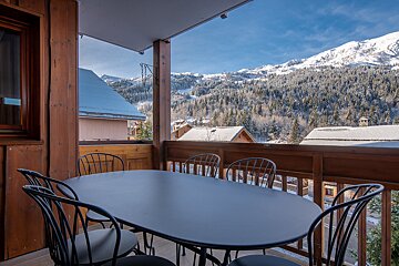 A wooden balcony with a dining table and chairs overlooks a stunning winter mountain landscape with snow-covered trees and distant chalets.