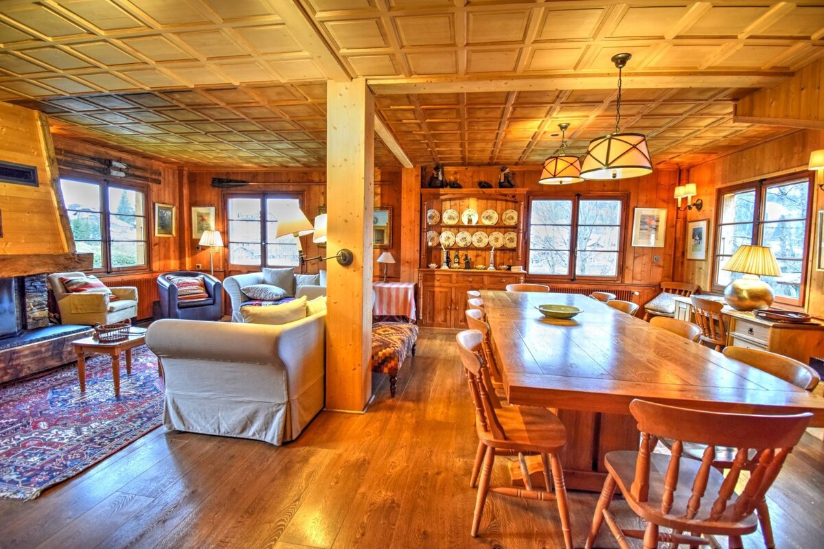 A warm, rustic open-plan living and dining room with extensive wood paneling, a large fireplace, comfortable seating, and a long dining table.