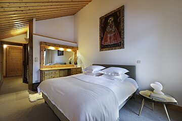 A cozy, rustic bedroom with a white bed, wooden slatted ceiling, and a large mirror with a dressing table. A traditional painting adorns the wall.