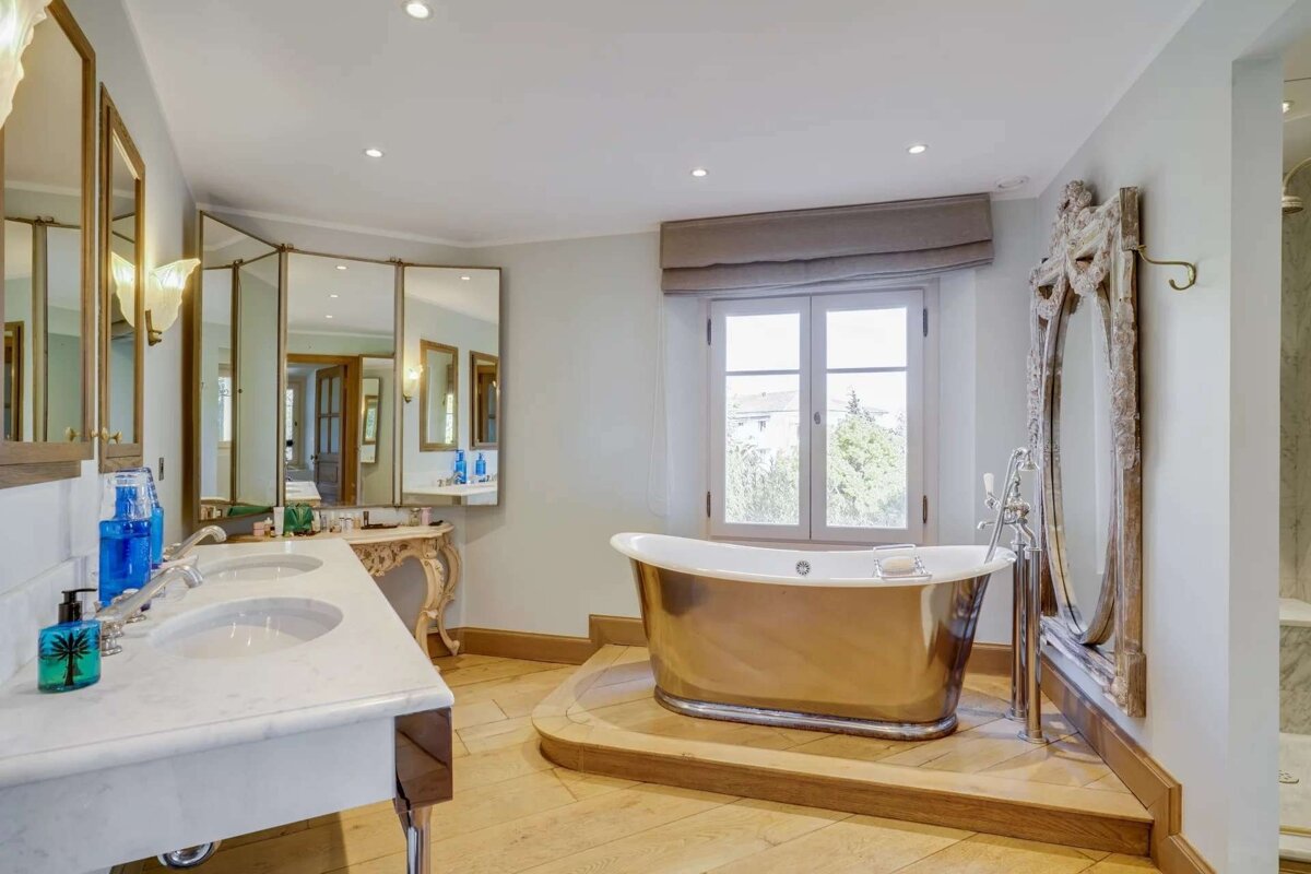 Elegant bathroom with a golden freestanding tub by a window, double sinks, ornate mirrors, and light wood flooring.