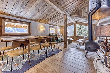 Rustic wood chalet interior with dining area, cozy lounge with stove, and large windows overlooking snowy mountains.