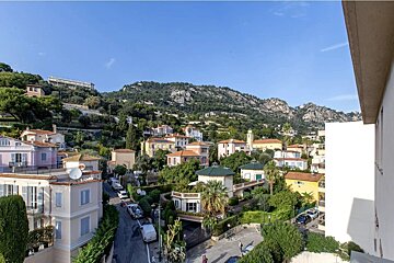 A picturesque hillside town with colourful houses nestled among green trees under a clear blue sky, with mountains in the background.