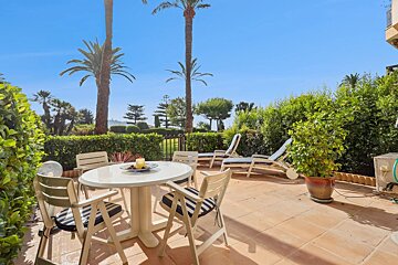 A sunny tiled patio with a dining table and chairs, lounge chairs, lush green hedges, and tall palm trees under a blue sky.