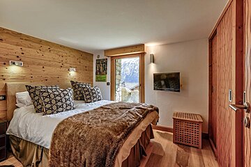 A cozy, modern bedroom features wood paneling, a large bed with a fur throw, a wall-mounted TV, and a window opening to a snowy mountain view.