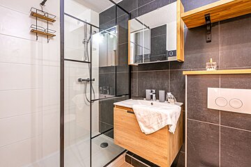 A modern bathroom featuring a glass shower, wooden vanity, mirror cabinet, and sleek dark gray and white tiled walls with wooden shelves.