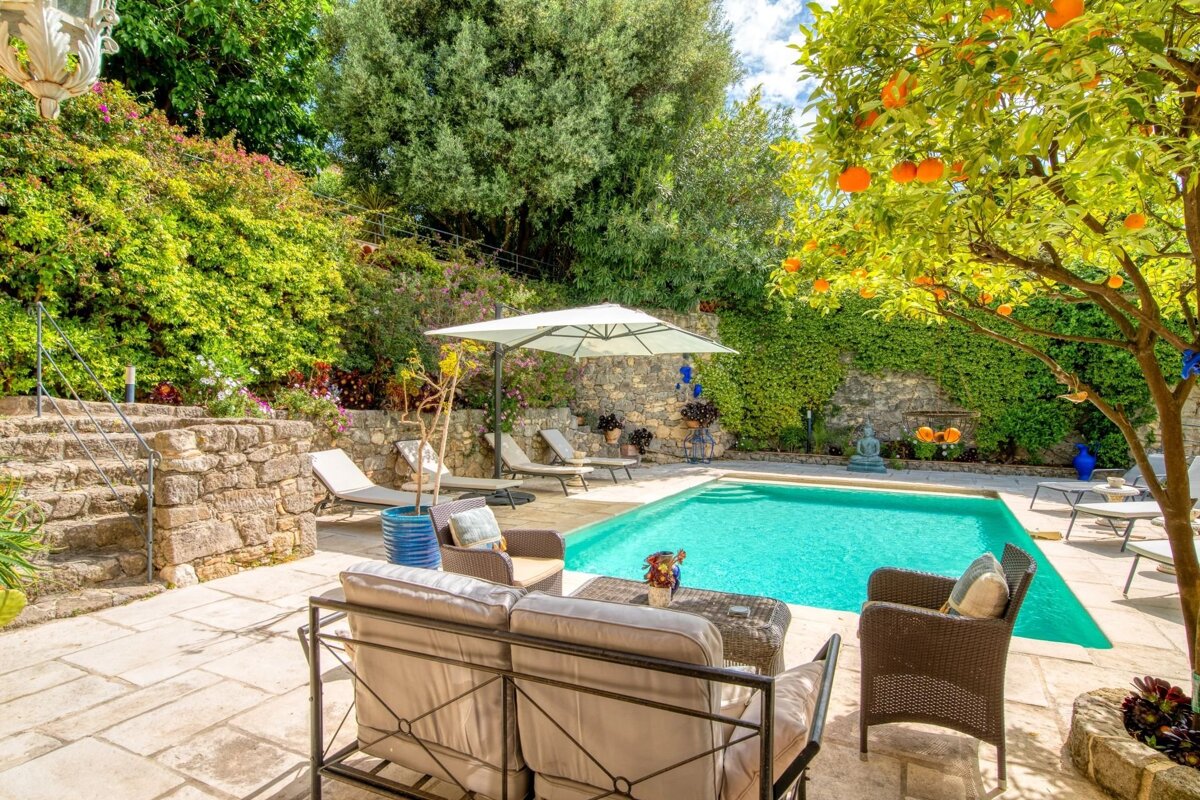 A sunny, lush outdoor patio with a bright blue swimming pool, comfortable lounge seating, and an orange tree bearing fruit.
