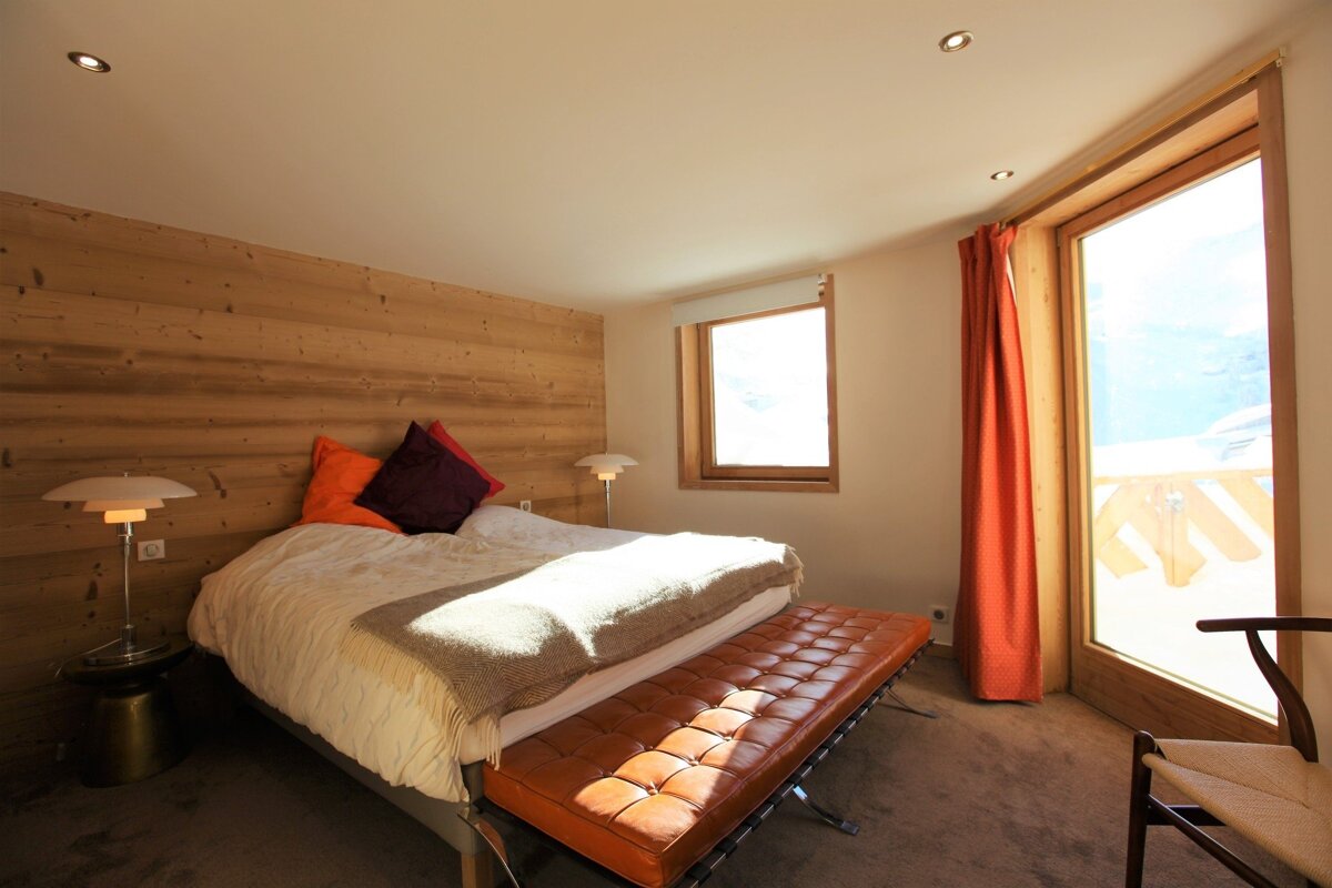 Cozy modern bedroom with a wood-paneled accent wall, a large bed, an orange tufted leather bench, and glass patio doors.