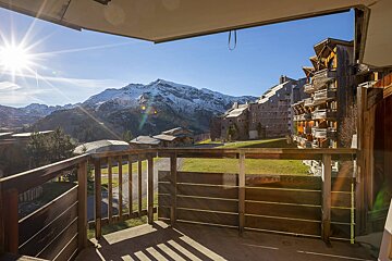 A sunny balcony view of snow-capped mountains, green slopes, and distinctive wooden alpine resort buildings under a clear blue sky.