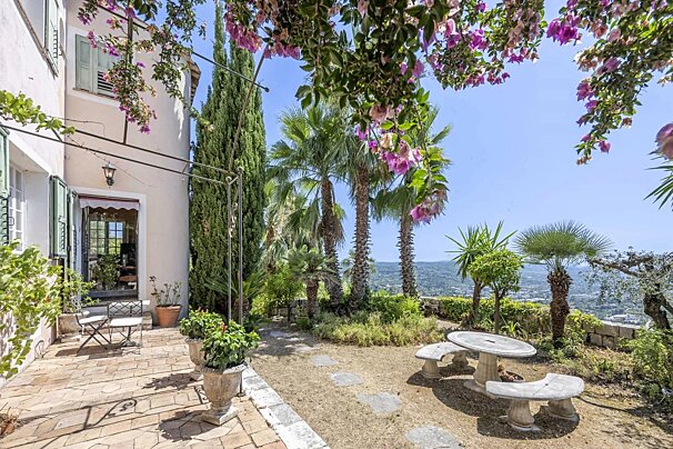 A sunny garden patio overlooking a valley, with pink bougainvillea, palm trees, and a stone table with benches.