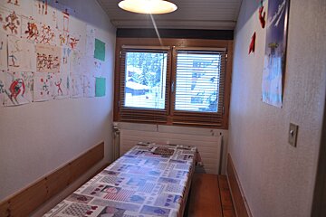 A narrow room features a single bed, a window with blinds showing a snowy view, and walls decorated with children's drawings and a poster.