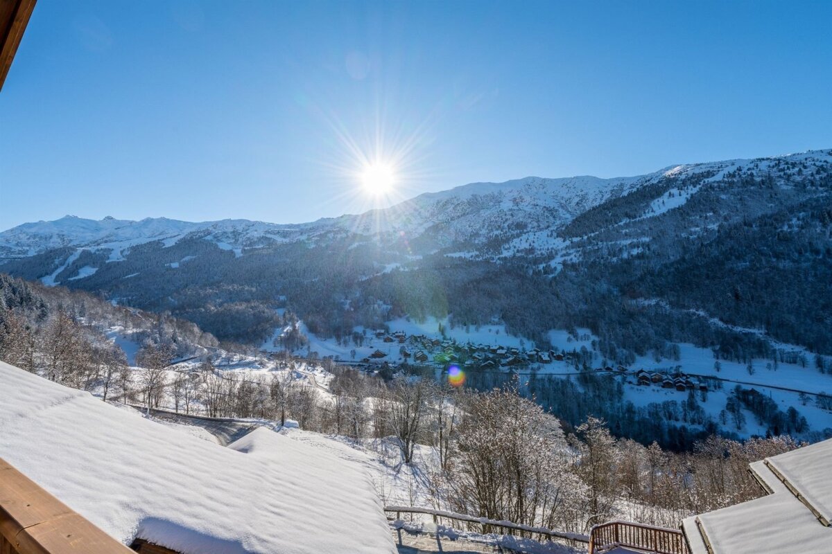 Sun-drenched winter mountain scene with snow-covered peaks, forests, and a village nestled in the valley beneath a clear blue sky.