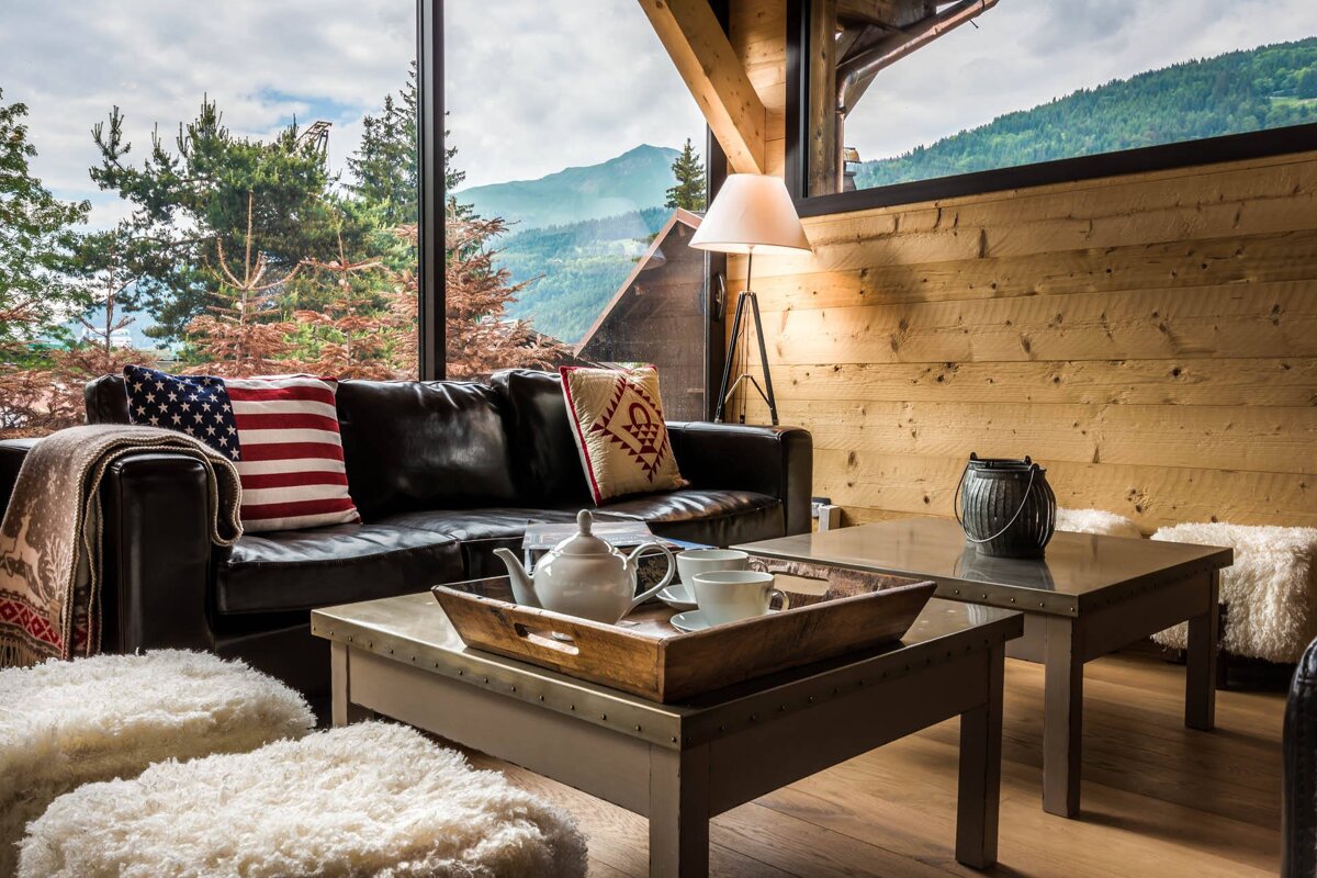 A cozy mountain lodge living room with a leather sofa, tea set, and large windows revealing a scenic mountain view. Features warm wood interior.
