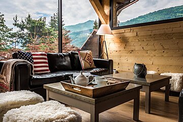 Cozy rustic living room with a leather sofa, tea set, fluffy stools, and a large window overlooking scenic mountains.
