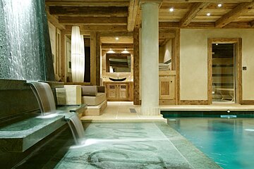 A luxurious indoor pool with a multi-tiered waterfall feature, exposed wooden beams, a vanity area, and a sauna, creating a rustic, spa-like ambiance.