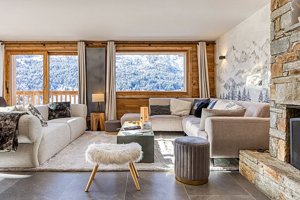 Cozy chalet living room with light sofas, a stone fireplace, wood paneling, and large windows overlooking snowy mountains and forests.