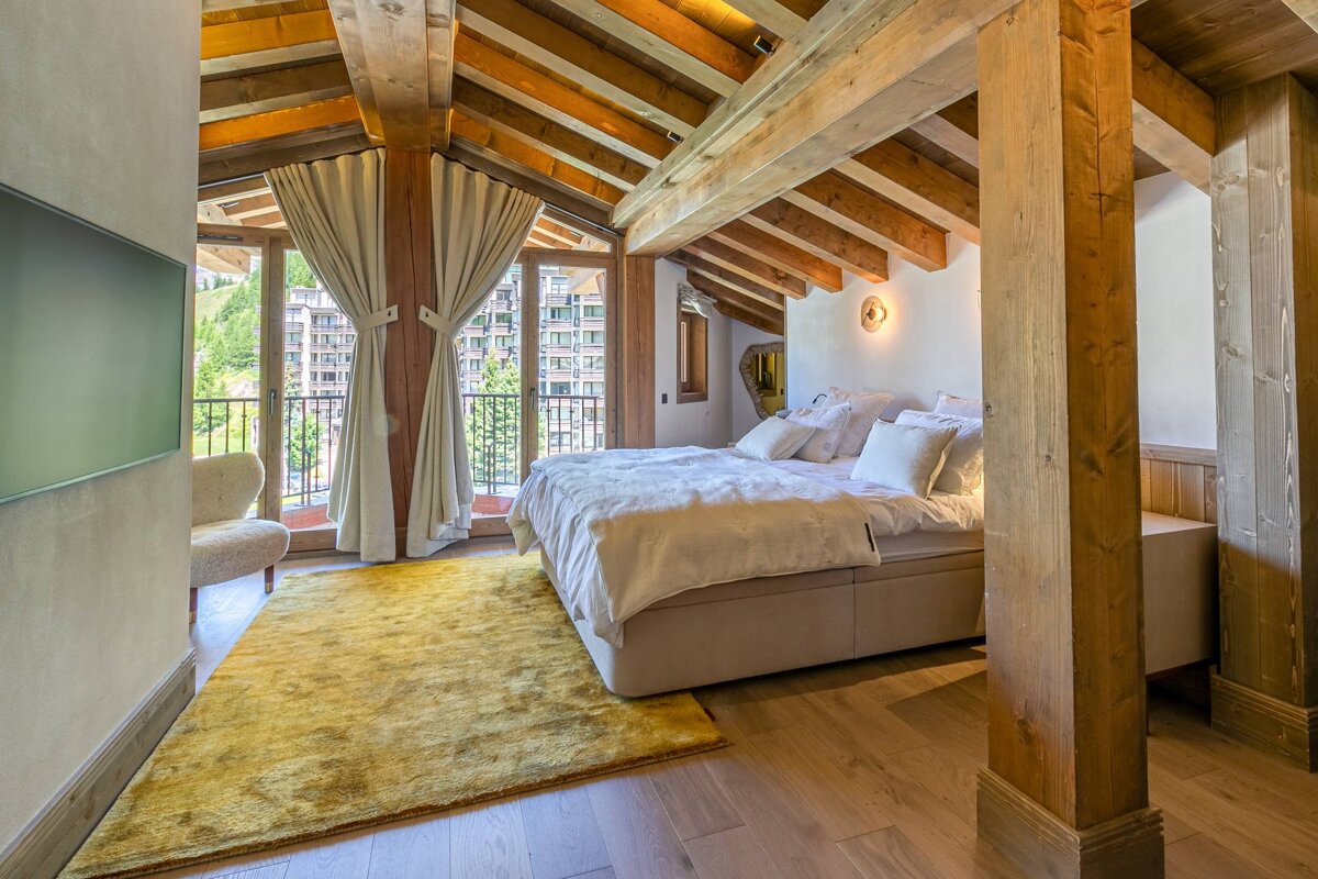 Stylish bedroom with exposed wooden beams, a comfy bed, bright yellow rug, and a balcony with mountain and building views.