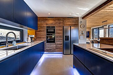 Modern kitchen featuring navy blue cabinets, wooden wall panels, stainless steel appliances, and cool underglow lighting.