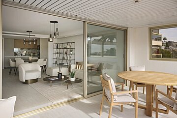 A balcony with a wooden dining set looks into a chic, modern living room through open sliding glass doors.