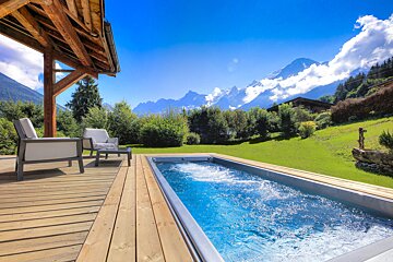 A luxurious wooden deck with seating and a bubbling hot tub overlooks a majestic, snow-capped mountain range under a bright blue sky.