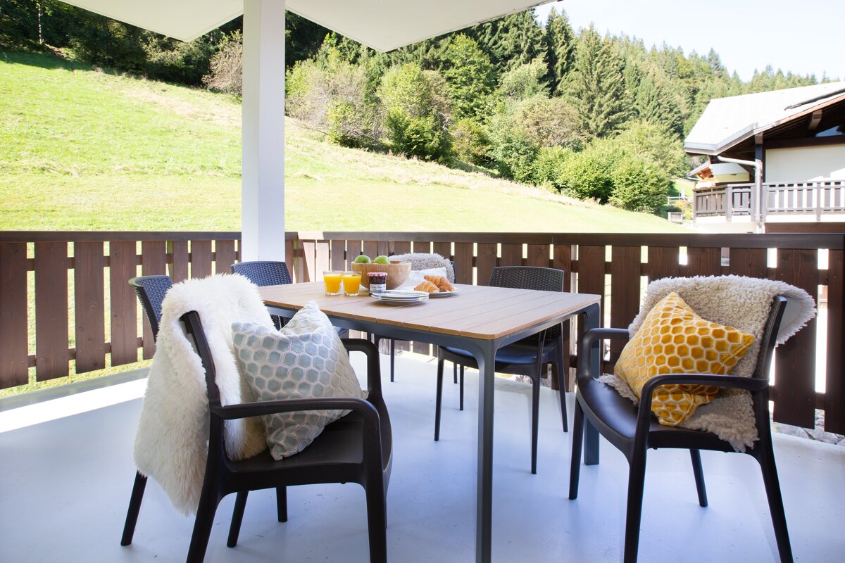 A sunny balcony with a table set for breakfast, cozy chairs with throws, and a view of a lush green mountain, forest, and distant house.