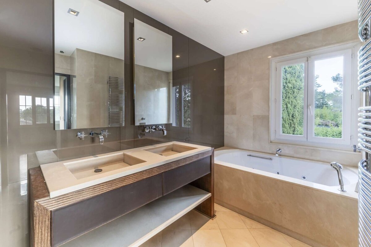 A modern bathroom features a double vanity with large mirrors, a built-in bathtub by a window with a green view, and neutral, earthy tones.