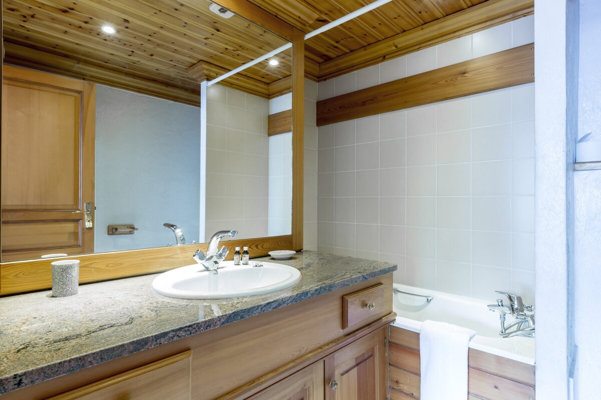 Chalet-style bathroom with wood-paneled ceiling & vanity, granite countertop, sink, large mirror, tiled walls, and a bathtub.