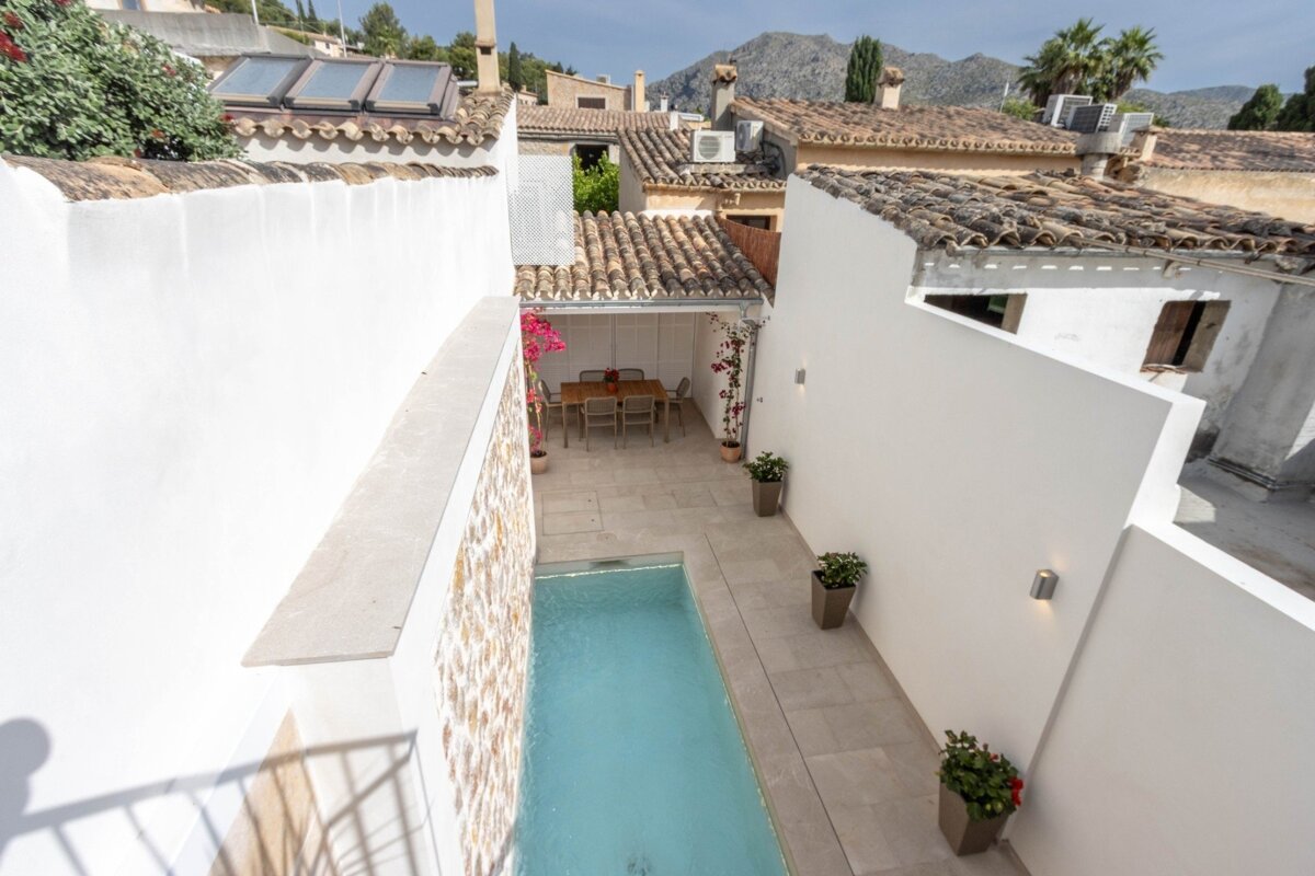 A high-angle view of a Mediterranean courtyard featuring a narrow pool, outdoor dining area, white walls, and terracotta roofs, with mountains in the distance.