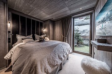 Cozy, wood-paneled bedroom with a plush bed, facing a window revealing a snowy winter landscape and a patch of green grass.