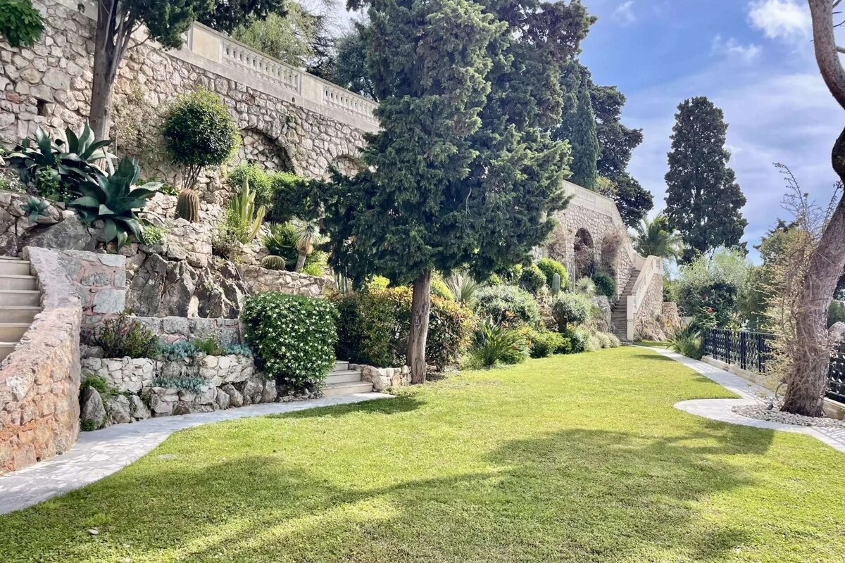 Sunlit terraced garden with stone walls, stairs, and vibrant Mediterranean foliage like cypress trees and cacti, alongside a manicured green lawn.