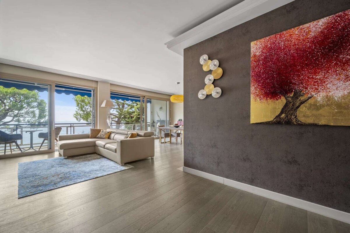 Bright modern living room with a cream sectional, ocean view balcony, and a striking red tree painting on a dark accent wall.