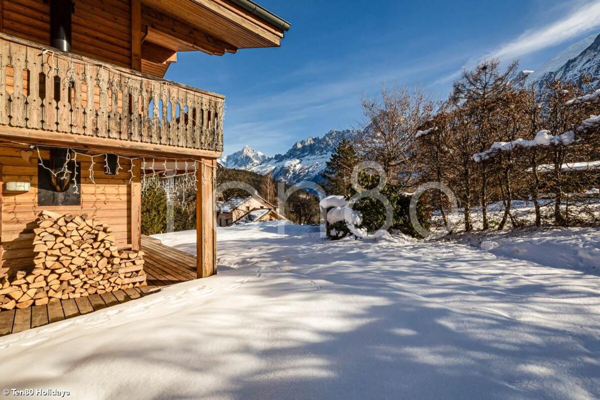 A pile of logs sits outside of a wooden cabin in the snow