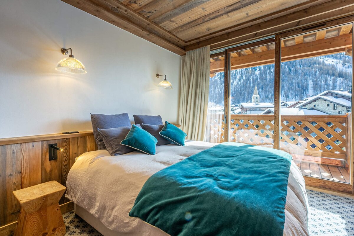 A cozy, rustic bedroom with a teal bed and wooden accents, featuring a balcony offering a stunning view of a snow-covered village and mountains.